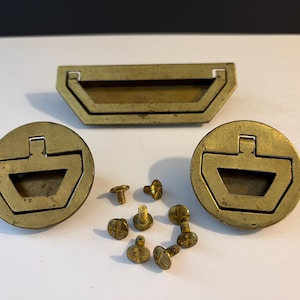 May include: A set of vintage brass hardware components, including a rectangular handle and two circular pulls. Several brass screws are also included. The hardware has a weathered appearance, suggesting age and use. Ideal for furniture restoration or DIY projects.