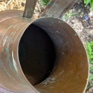 May include: A copper watering can with a worn, aged patina. The can has a wide, round opening and a handle that curves up and over the top.