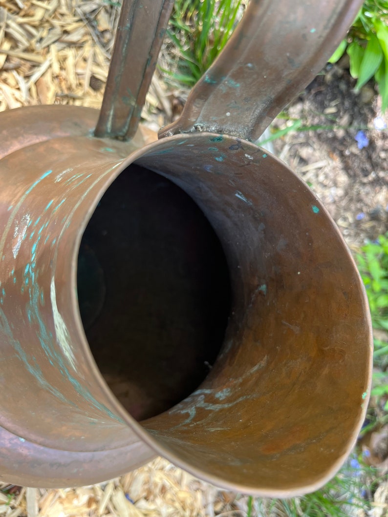 May include: A copper watering can with a worn, aged patina. The can has a wide, round opening and a handle that curves up and over the top.