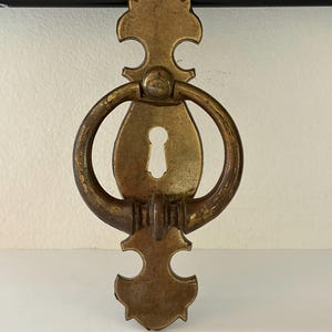 May include: Antique brass drawer pull with a keyhole and a decorative design. The pull features a ring handle and ornate top and bottom details. The hardware is mounted on a white surface.