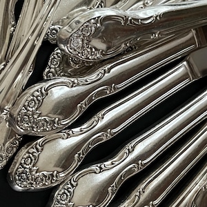 May include: Close-up of a collection of ornate silverware. The silver utensils, including knives and spoons, feature detailed floral patterns on the handles. The reflective surfaces create highlights and shadows, emphasizing the intricate designs.