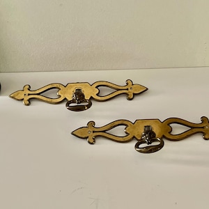 May include: Two ornate, brass-toned drawer pulls. Each features a decorative, elongated design with a central ring pull and pointed ends. The hardware is likely for furniture restoration or adding a vintage touch.