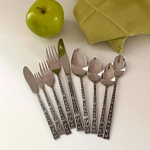 May include: A set of stainless steel silverware with decorative handles. The set includes knives, forks, and spoons. A green apple and a folded green napkin are also in the image.