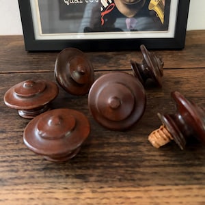 May include: Six dark brown bottle stoppers with a tiered, circular design. One stopper has a cork base. The stoppers are arranged on a dark wooden surface, with a framed picture in the background.