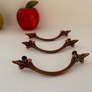 May include: Three antique bronze-colored drawer pulls with a curved design and decorative ends. The pulls are arranged on a white surface, with a red apple and a yellow pear in the background.