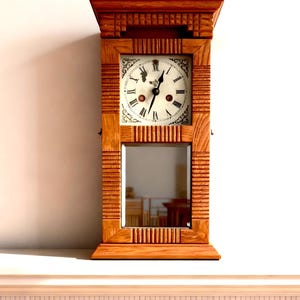 May include: A wooden wall clock with a rectangular frame and a mirrored bottom section. The clock face is white with black Roman numerals and ornate hands. The wood has a warm, reddish-brown tone, and the clock is mounted on a white wall.