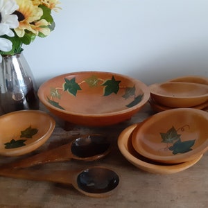 May include: A collection of wooden bowls and serving spoons. The bowls are various sizes, with a warm orange hue and green leaf designs. Two large wooden spoons with dark brown bowls are also present. A vase of flowers is in the background.