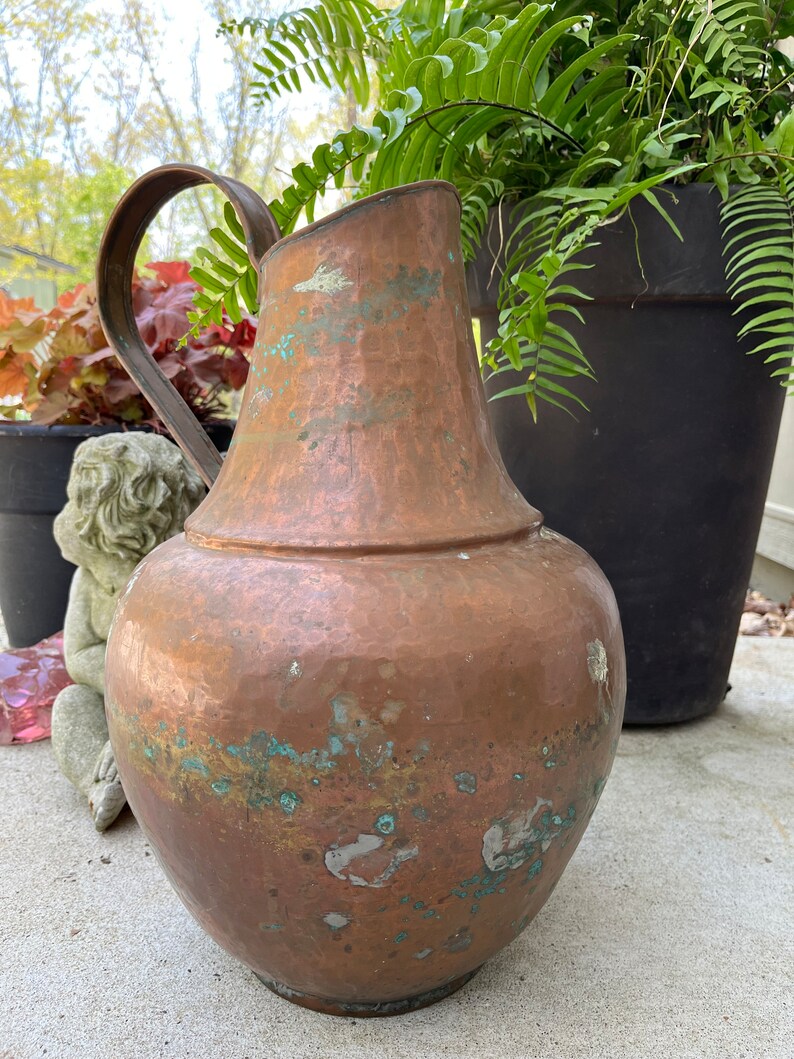 May include: A large, hammered copper pitcher with a wide mouth and a curved handle. The pitcher has a patina of age and shows signs of wear, with green and brown spots on the surface.