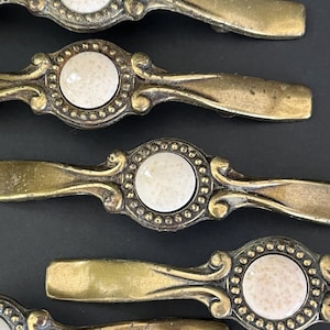 May include: Close-up of several antique brass cabinet pulls. Each pull features a central, round, off-white ceramic insert surrounded by a decorative brass border. The pulls have a curved handle design, suggesting a vintage aesthetic.
