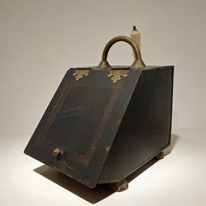May include: A black metal coal scuttle with a brass handle and decorative accents. The scuttle has a hinged front panel and sits on small, claw-shaped feet. A candle is placed on top of the scuttle.