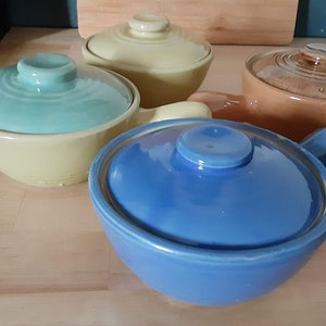 May include: Four ceramic bowls with lids in various colors: blue, green, yellow, and orange. Each bowl has a handle and a matching lid with a knob. The bowls are arranged on a light-colored wooden surface.
