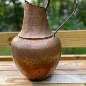 May include: A hammered copper pitcher with a wide mouth and a curved handle. The pitcher has a rustic, aged look and is made of a thick, durable metal.