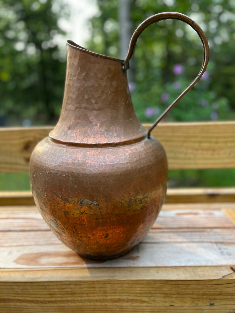 May include: A hammered copper pitcher with a wide mouth and a curved handle. The pitcher has a rustic, aged look and is made of a thick, durable metal.