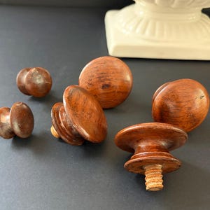 May include: A collection of six round wooden knobs with a polished brown finish. The knobs vary in size and shape, featuring smooth, rounded tops and cylindrical bases. Some knobs show visible wood grain patterns and threaded metal or wooden screws.