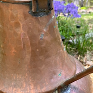 May include: A large, antique copper pitcher with a hammered finish and a worn handle. The pitcher has a patina of age and shows signs of use.