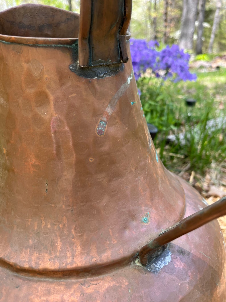 May include: A large, antique copper pitcher with a hammered finish and a worn handle. The pitcher has a patina of age and shows signs of use.