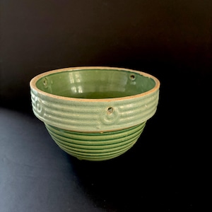 May include: A sage green ceramic bowl featuring a ribbed texture on the exterior and a smooth interior. The upper rim is accented with a series of small, evenly spaced circular indentations, and the base has a rounded, tapered shape.