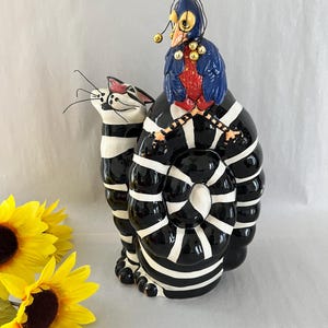 May include: A black and white striped ceramic cat figurine with a blue and red bird perched on its back. The bird has gold bells hanging from its head.