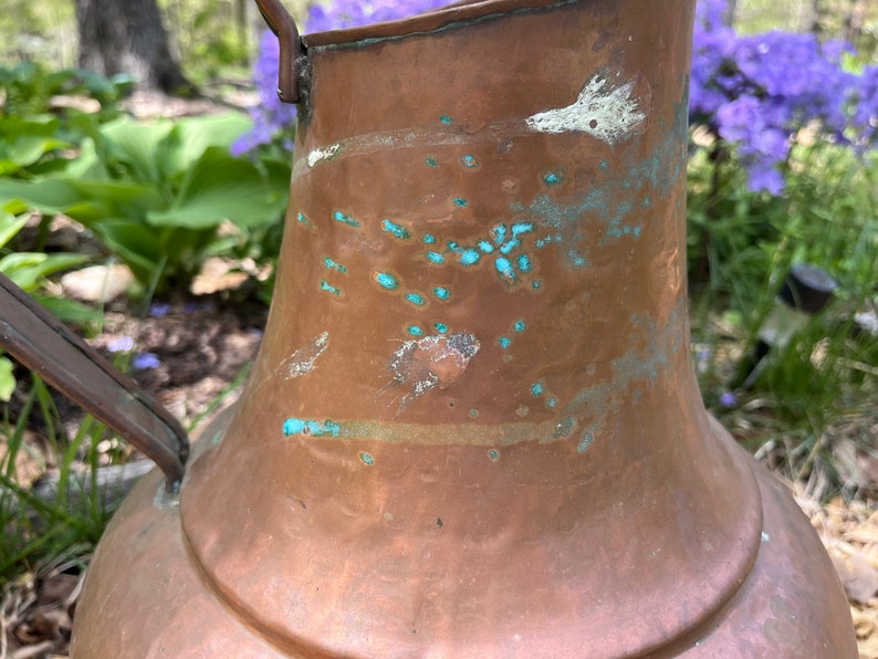 May include: A large copper pitcher with a handle. The pitcher has a patina of green and white spots.