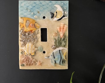May include: Decorative light switch cover featuring an underwater scene with colorful fish, coral, and a net design. The cover is rectangular with a single switch opening and two screw holes. The color palette includes shades of blue, yellow, and beige.
