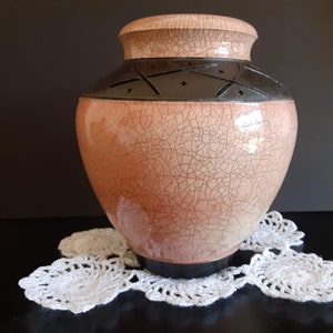 May include: A pink ceramic vase with a crackled glaze and a black band around the top. The band has a decorative design of stars and crosses.