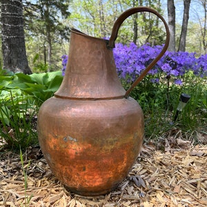 May include: A large, copper pitcher with a hammered finish and a rounded body. The pitcher has a single handle and a wide mouth. It is sitting on a bed of wood chips in a garden setting.