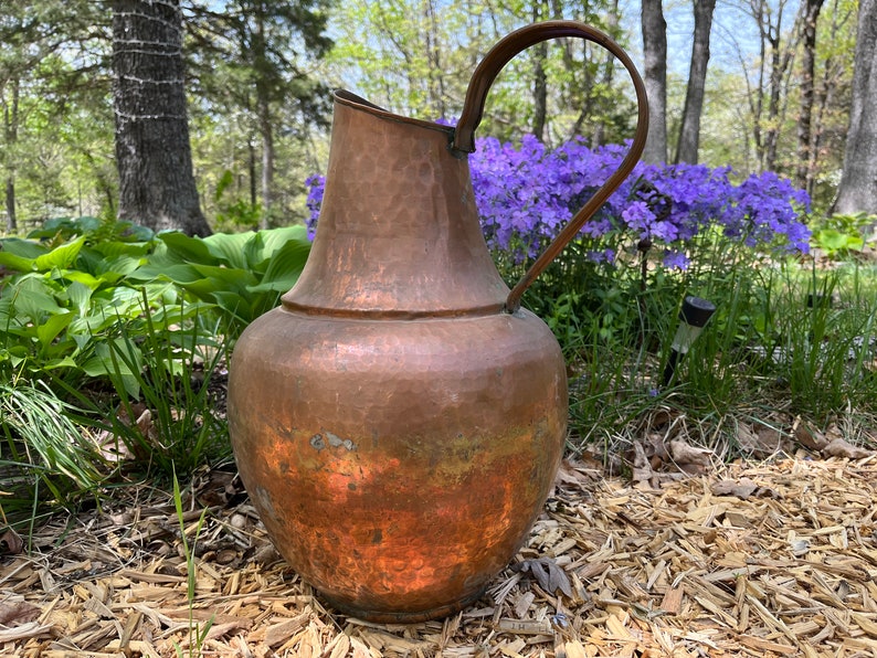 May include: A large, copper pitcher with a hammered finish and a rounded body. The pitcher has a single handle and a wide mouth. It is sitting on a bed of wood chips in a garden setting.