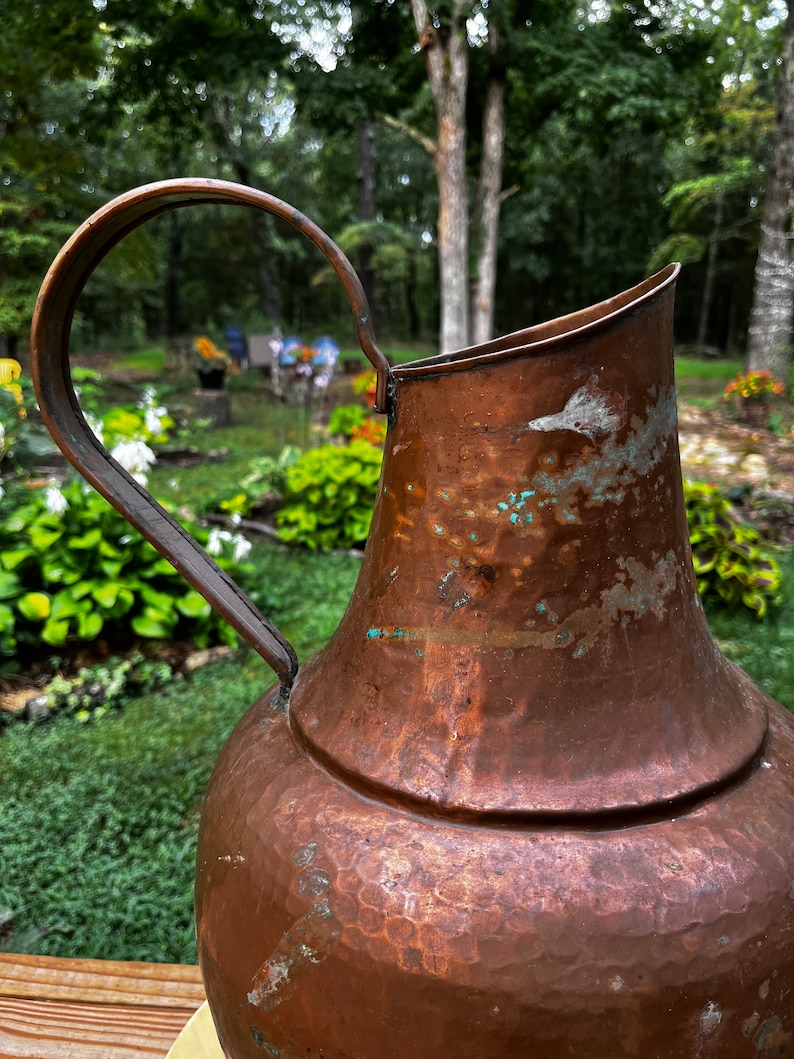 May include: A large, copper pitcher with a hammered finish. The pitcher has a wide mouth and a handle that curves up and over the top. The pitcher is slightly tarnished, giving it a vintage look.