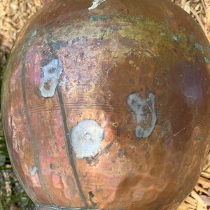 May include: A large copper pot with a rounded body and a narrow neck. The pot has a worn, aged appearance with visible scratches and marks. The pot is sitting on a bed of straw.