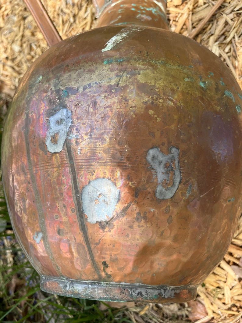 May include: A large copper pot with a rounded body and a narrow neck. The pot has a worn, aged appearance with visible scratches and marks. The pot is sitting on a bed of straw.