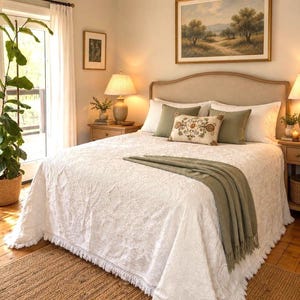May include: A white bedspread with a textured floral pattern and fringed edges, featuring a sage green throw blanket, two sage green pillows, and a rectangular accent pillow with a colorful embroidered floral design on a bed.