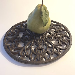 May include: A green pear with a leaf rests on a decorative, dark brown, cast iron trivet. The trivet is round with an ornate, openwork design. The pear is light green with a brown stem.