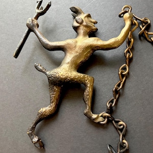 May include: A metallic bronze-toned wall ornament shaped like a horned figure with a tail, holding a three-pronged pitchfork in one hand and attached to a metal chain that hangs from the other hand, featuring a textured, weathered finish.