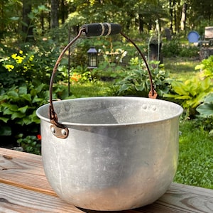 May include: A vintage, aluminum bucket with a curved handle and a black and white grip. The bucket has a rounded shape and is set on a wooden surface, with a natural background of green foliage and trees.
