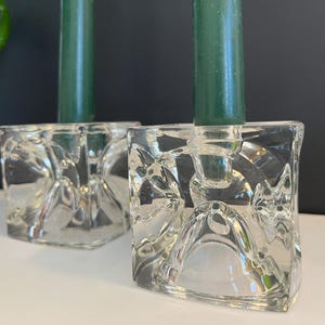 May include: Two clear glass candle holders with green candles. The candle holders are square with a textured, geometric design. The candles are a deep green color, and the background is a mix of black and white.