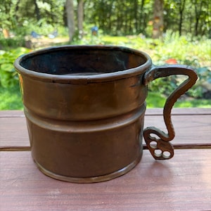 May include: A cylindrical copper mug with a dark, weathered patina and a horizontal ridge around the center. It features a distinct, textured metal handle with a decorative, open-work scroll design at the base, showing signs of oxidation.