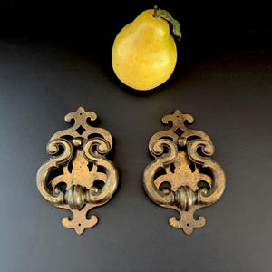 May include: Two matching antique brass door knockers featuring an ornate, symmetrical scrollwork design with a central swinging handle. The metal has a darkened, aged patina finish. A single yellow pear is positioned above the two door knockers.