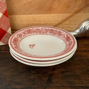 May include: A stack of three white ceramic plates with a red floral border design. The plates have a shallow bowl shape and a small logo in the center. The plates are on a dark wooden surface.