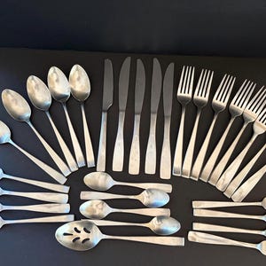 May include: A large collection of stainless steel flatware including various spoons, dinner knives, and forks. The set features a simple, smooth design with rounded handles. The pieces are arranged in a grid-like pattern on a dark surface.