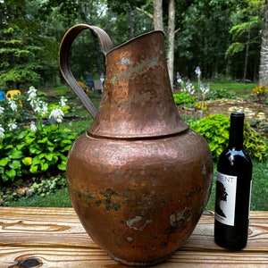 May include: A large, hammered copper pitcher with a handle sits on a wooden table next to a bottle of red wine. The pitcher has a rustic, aged look and is a beautiful example of vintage home decor.