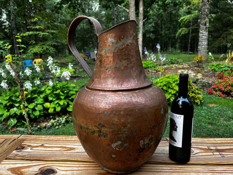 May include: A large, hammered copper pitcher with a handle sits on a wooden table next to a bottle of red wine. The pitcher has a rustic, aged look and is a beautiful example of vintage home decor.