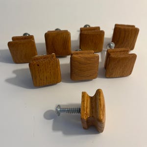 May include: A collection of eight wooden cabinet knobs with a warm, medium-brown finish. Each knob has a rectangular shape with rounded edges and a silver-colored metal screw. The knobs are arranged on a white surface.