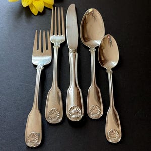 May include: A five-piece silver-toned flatware set consisting of two forks, one knife, and two spoons of varying sizes. Each handle features a distinct embossed shell motif at the base, providing a uniform look across the entire cutlery collection.