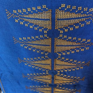 以下が含まれることがあります： 鮮やかな青い生地に、黄金色の糸で刺繍された左右対称の様式化された葉のような模様が縦に並んでいます。刺繍は緻密なクロスステッチのディテールで構成されており、質感のある繰り返しデザインを作り出しています。