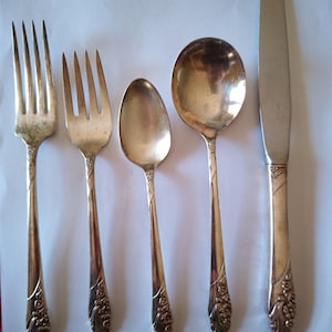 Puede incluir: Un juego de cubiertos antiguos, que incluye dos tenedores, dos cucharas y un cuchillo. La cubertería plateada presenta detalles florales ornamentados en los mangos. Los utensilios están dispuestos en fila sobre un fondo claro, mostrando su diseño clásico.