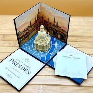 Könnte beinhalten: Eine Pop-up-Grußkarte mit einem dreidimensionalen Papiermodell der Dresdner Frauenkirche. Das Innere zeigt ein gedrucktes Stadtbild von Dresden bei Sonnenuntergang. Der Text auf der Karte lautet: EINE REISE NACH DRESDEN, Zum Verschenken & Erinnern und REISEGUTSCHEIN.