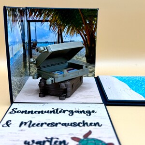 Könnte beinhalten: Eine schwarz-weiße Explosionsbox mit einem Miniatur-Koffer, einer Strandszene und dem Text "Sonnenuntergänge & Meeresrauschen warten".