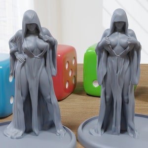 Puede incluir: Dos figuritas grises pintadas de una persona con una túnica con capucha. Las figuritas están de pie sobre bases redondas. Las bases están sobre una superficie de madera. Hay cuatro dados en el fondo.