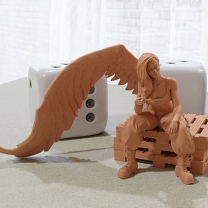 May include: A brown figurine of a woman with wings sitting on a wooden pallet. The woman is wearing a sleeveless top and pants. Her wings are spread out behind her.