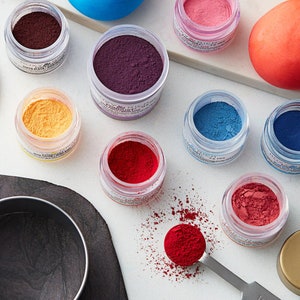 May include: Assortment of small glass jars filled with colorful powder pigments. The jars are arranged on a white surface with a black slate tile in the background. The pigments are red, orange, yellow, blue, purple, and pink.